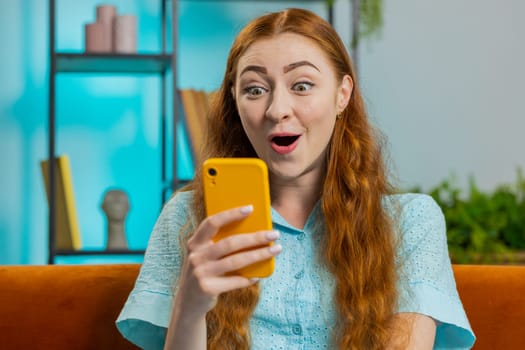 Happy young woman use mobile smartphone typing browsing say Wow yes found out great big win good news celebrate lottery jackpot doing winner gesture. Redhead girl at home in room sitting on sofa