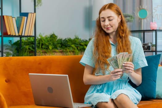 Planning budget. Rich happy redhead woman counting money cash use laptop computer calculate domestic bills at home. Joyful Girl satisfied of income earnings, saves money for planned vacation, gifts