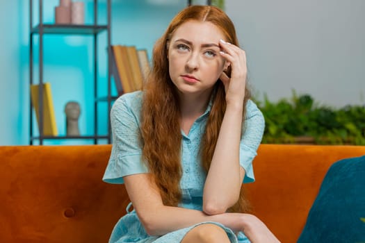 Sad lonely redhead woman sits at home room looks pensive thinks over life concerns or unrequited love suffers from unfair situation. Alone girl problem break up depressed feeling bad annoyed burnout