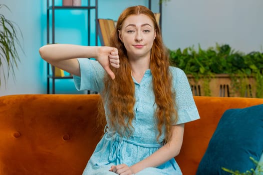 Dislike. Portrait of upset redhead woman showing thumbs down sign gesture, expressing discontent, disapproval dissatisfied bad work at modern home apartment indoors. Displeased girl in room on sofa