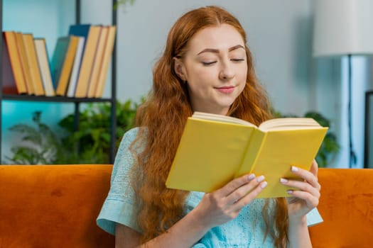 Young redhead woman relaxing interesting book, turning pages smiling enjoying literature, taking a rest on comfortable couch. Portrait of peaceful cheerful girl at home apartment living room on sofa
