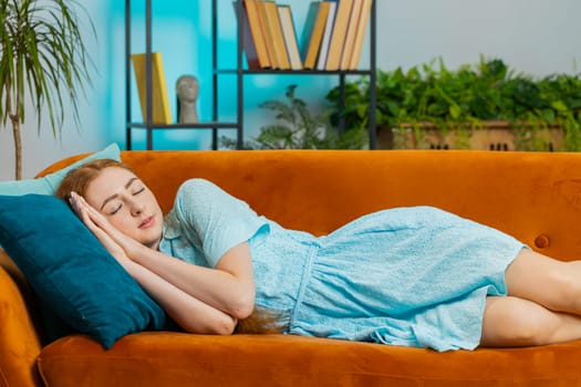 Tired young redhead woman lying down in bed taking a rest at home. Portrait of carefree girl napping, falling asleep on comfortable orange sofa with pillows. Closed her eyes enjoy daytime nap alone