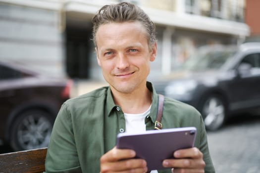 Texting message concept as man smiles while holding tablet PC on bench outdoors. With joyful expression on face, he engages in digital communication and stays connected in urban environment. High quality photo