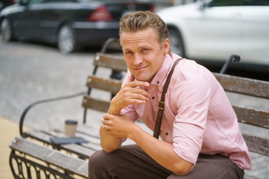 Handsome individual seated on a bench in the charming streets of an old town. With a warm and genuine smile, he confidently gazes at the camera, radiating charm and positive energy. . High quality photo