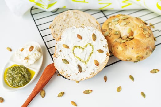A freshly baked bagel with Philadelphia cheese, sunflower seeds scattered on the table and a heart painted with pesto sauce. Delicious and healthy breakfast