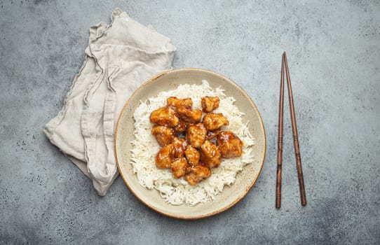 Chinese sweet and sour sticky chicken with sesame seeds and rice on ceramic plate with chopsticks top view on gray rustic stone background, traditional dish of China
