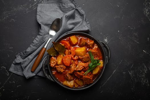 Beef meat stew with potatoes, carrot and delicious gravy in black casserole pot with bay leaves with spoon on black dark rustic concrete background from above .