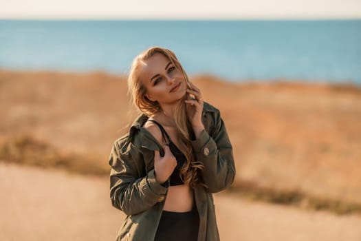 Portrait blonde sea cape. A calm young blonde in an unbuttoned khaki raincoat stands on the seashore, under the raincoat there is a black skirt and top.