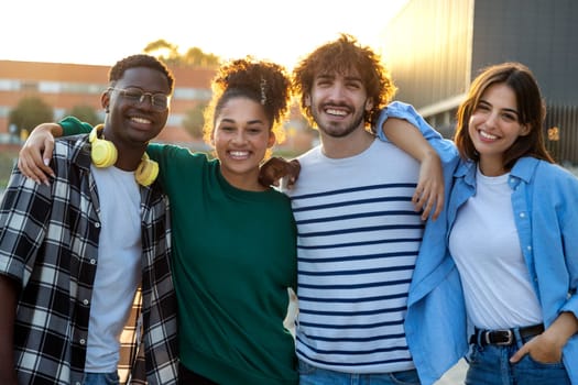 Group of happy and smiling multiracial friends embracing looking at camera standing outdoors during sunset. Youth lifestyle concept.