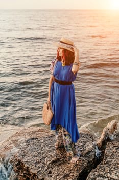 A woman in a dress, hat and with a straw bag is standing on the beach enjoying the sea. Happy summer holidays.