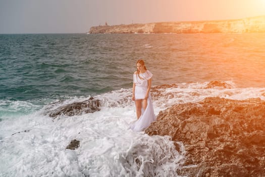 A woman stands on a rock in the sea during a storm. Dressed in a white long dress, the waves break on the rocks and white spray rises