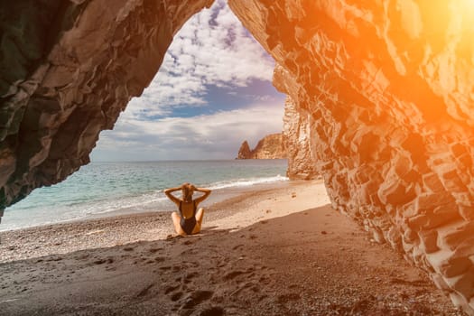Woman travel sea. View of a woman in a black swimsuit from a sea cave Attractive woman enjoying the sea air sits on the beach and looks at the sea. Behind her are rocks and the sea.