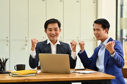 Happy business people looking at laptop screen excited by good news online, celebrating success raising hands up.