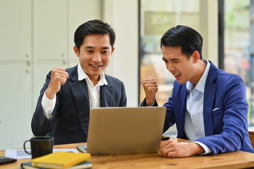 Happy business people looking at laptop screen excited by good news online, celebrating success raising hands up.