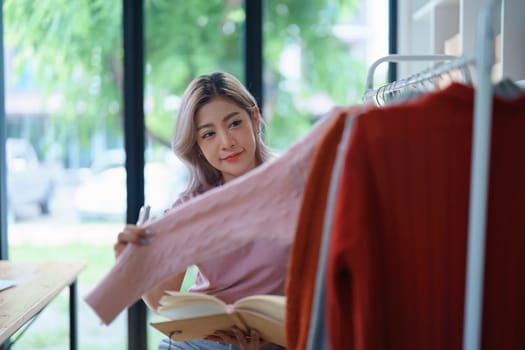 Portrait Of Asian Female Owner Of Fashion Store Checking Stock In Clothing Store With using notebook successful happy smile at small business, sme or ecommerce concepts.