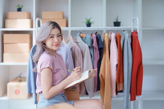 Portrait Of Asian Female Owner Of Fashion Store Checking Stock In Clothing Store With using notebook successful happy smile at small business, sme or ecommerce concepts.