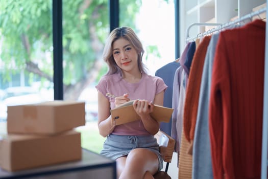 Portrait Of Asian Female Owner Of Fashion Store Checking Stock In Clothing Store With using notebook successful happy smile at small business, sme or ecommerce concepts.