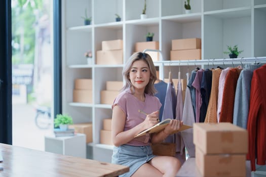 Portrait Of Asian Female Owner Of Fashion Store Checking Stock In Clothing Store With using notebook successful happy smile at small business, sme or ecommerce concepts.