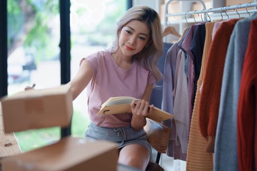 Portrait Of Asian Female Owner Of Fashion Store Checking Stock In Clothing Store With using notebook successful happy smile at small business, sme or ecommerce concepts.