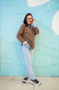 Happy woman in glasses outdoor on blue color background. Positive people concept. Smiling girl looking at camera, hands in pocket, dressed sweater and jeans. Vertical full length
