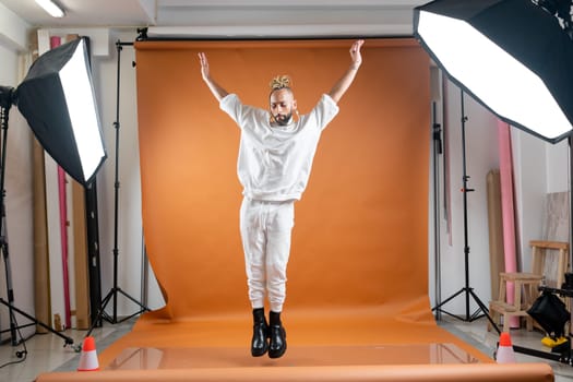 Full length african-american gay man with bright make up jumping wearing white dancing on orange studio background. Brazilian male posing in photo studio. Bearded afro-american gay man dancer