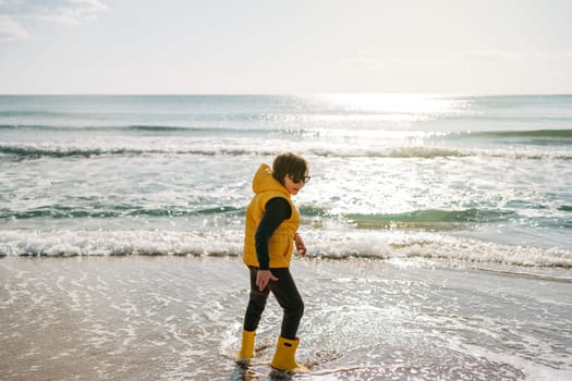 Boy in yellow rubber boots playing with sand at the beach. School kid touching water at autumn winter sea. Child having fun and jumping with waves at the shore. Spring Holiday vacation concept.