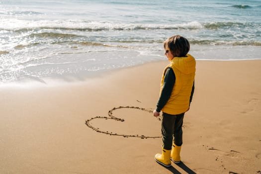 Boy in yellow rubber boots drawing heart shape on sand at the beach. School kid touching water at autumn winter sea. Child having fun with waves at the shore. Spring Holiday vacation concept.
