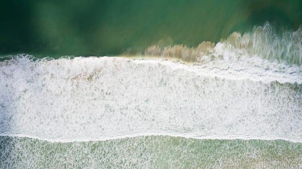 Top view of the stormy ocean and the shadows from the clouds. The waves raise the brown sand giving the emerald color of the water to the ocean. White foam. Cloud shadows create a dark gradient.