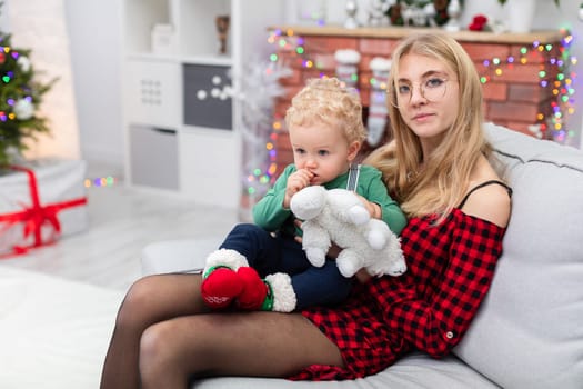 The woman is wearing an elegant plaid dress. The woman is holding a small child on her lap. Both are sitting on a gray couch. The room is decorated with Christmas decorations. The woman is wearing corrective glasses.
