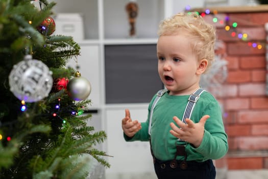 A little boy stands by a decorated Christmas tree. The child delights in the ornaments hung on the Christmas tree. Close-up on the figure of the child. Blurred background. in the background a brick fireplace decorated with colorful lights.