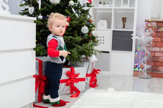 The child stands by the Christmas tree, under which are hidden large boxes of gifts. The boxes are tied with red ribbons. The Christmas tree is decorated. There are many Christmas decorations in the room.