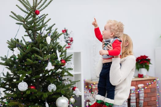 Mom holds her little son in her arms. The boy points to the top of the Christmas tree with his finger. The woman lifts the baby high into the air. Mom and son hang out by the Christmas tree.