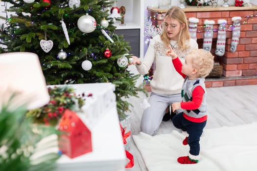A woman in a beige sweater wearing glasses kneels by a Christmas tree. Next to her stands a little boy. The family is looking for gifts hidden under the Christmas tree. Mother and son spend Christmas together.