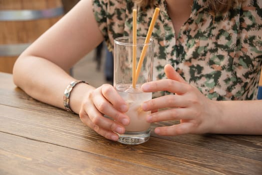 Cropped close-up of unrecognizable young woman holding glass with cold exotic cocktail, sitting at table in street cafe on sunny gold time, woman drinking cool lemonade. High quality photo