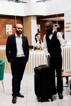 Bellboy leading businessman to room, carrying luggage for him and ensuring pleasant stay at hotel. Bellhop offering luxury concierge services to guest travelling for work, front desk.
