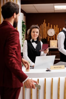 Employee checking guest passport to validate online booking reservation, man travelling for work. Front desk staff verifying identification documents and providing excellent concierge services.