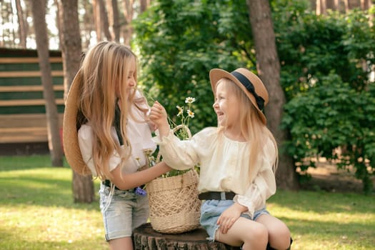 Two cheerful preteen girls sitting on tree stump and sniffing daisies culled while walking in green park. Happy sisters enjoying summer vacation