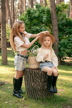 Smiling caring preteen girl adjusting hat to her little sister sitting on tree stump while walking together with in green sunny summer forest picking wildflowers