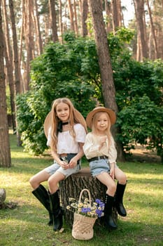 Two cheerful preteen girls sitting on big stump among tall trees, wicker basket full of fresh picked wildflowers standing on ground. Happy sisters resting during summer forest walk on sunny day