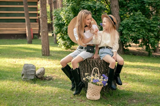 Two friendly preteen sisters wearing country style clothes enjoying walk in green summer park, sitting on tree stump and sniffing flowers. Happy childhood concept