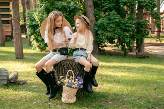 Two happy preteen sisters in light blouses, denim shorts, high boots and straw hats chatting merrily sitting on tree stump with colorful wildflowers in wicker basket nearby in green summer park