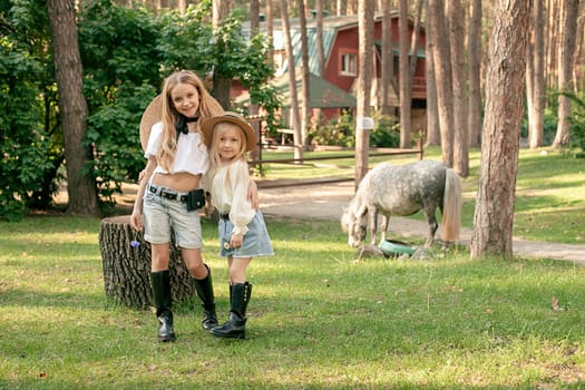 Cheerful preteen sisters enjoying spending time together on country estate in pine forest on sunny summer day, hugging while posing on lawn between tall trees on background of small gray pony