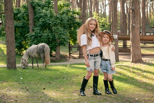 Two happy tween girls standing on green lawn on pine park on background of grazing dapple gray pony. Portrait of two smiling sisters hugging while walking. Sisterly affection concept