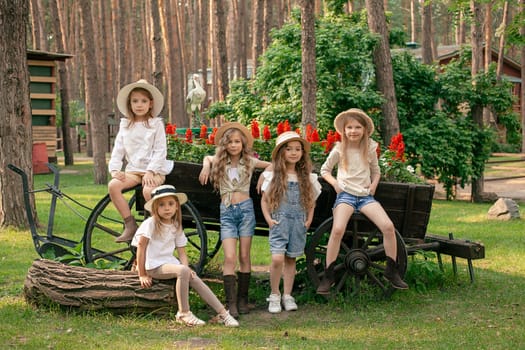 Group of friendly cute preteen girls in country style clothes posing together next to old vintage wooden cart designed as flower bed on green lawn in backyard of summer estate located in pine forest