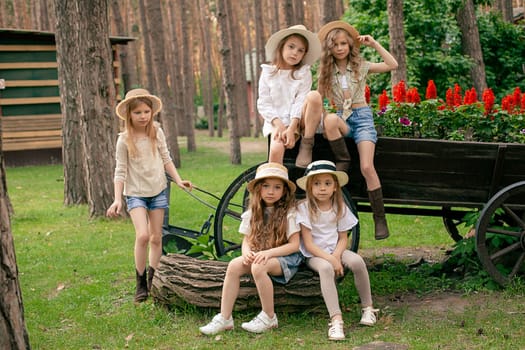 Group portrait of preteen girls in casual summer clothes and wicker sun hats walking in green city park, posing near old rustic wooden cart used as flower bed with blooming red salvia