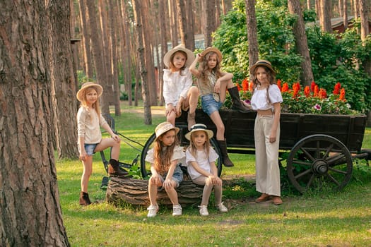 Group of cheerful preteen girls walking in landscaped summer park, posing together near decorative flower bed in shape of vintage wooden cart with blooming red flower