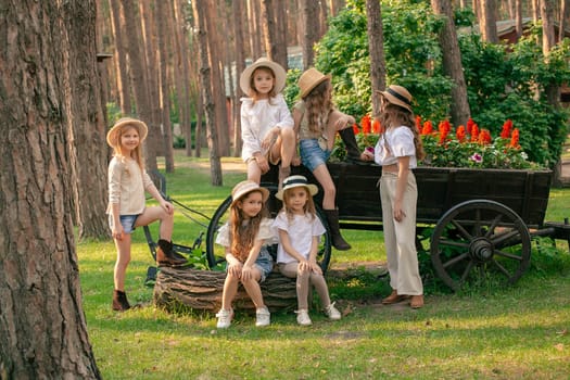 Portrait of friendly tween girls enjoying summer vacation at country estate posing on green lawn next to decorative wooden cart-shaped flower bed with blooming red salvia on sunny day.