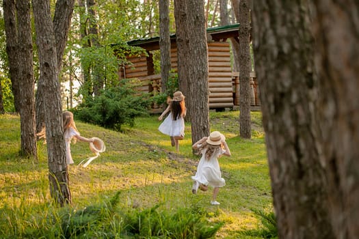 Rear view of preteen girls in light dresses and wicker hats having fun in country estate on sunny summer day, playing tag running among tall trees on green lawn