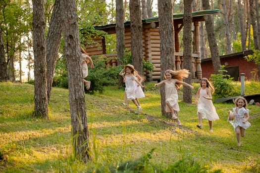 Group of happy preteen girls in light dresses having fun in country estate on sunny summer day, playing tag running among tall trees on green lawn