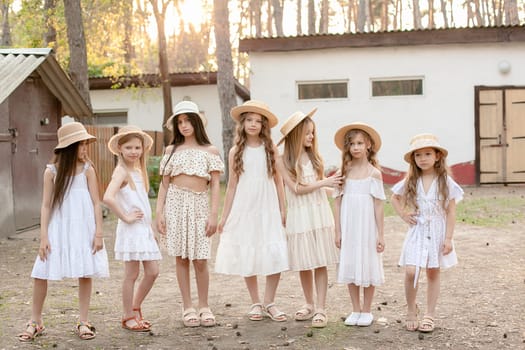 Group of cute preteen girls in summer dresses and wicker hats posing together on backyard of country estate surrounded by tall trees on sunny day
